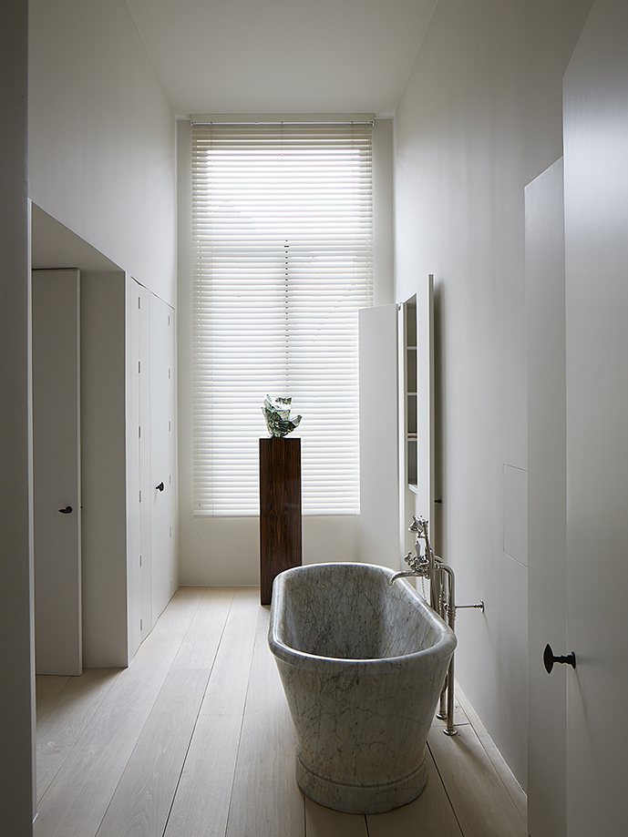 A monolithic white marble bathtub sits on pale floorboards beneath a tall blind-shaded window, with chrome fittings and flush white doors along the walls.