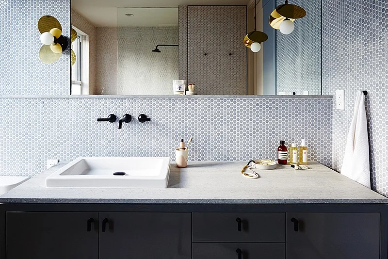 A stone-topped vanity with black pulls and brass sconces sits in front of mirrored walls and penny tile.