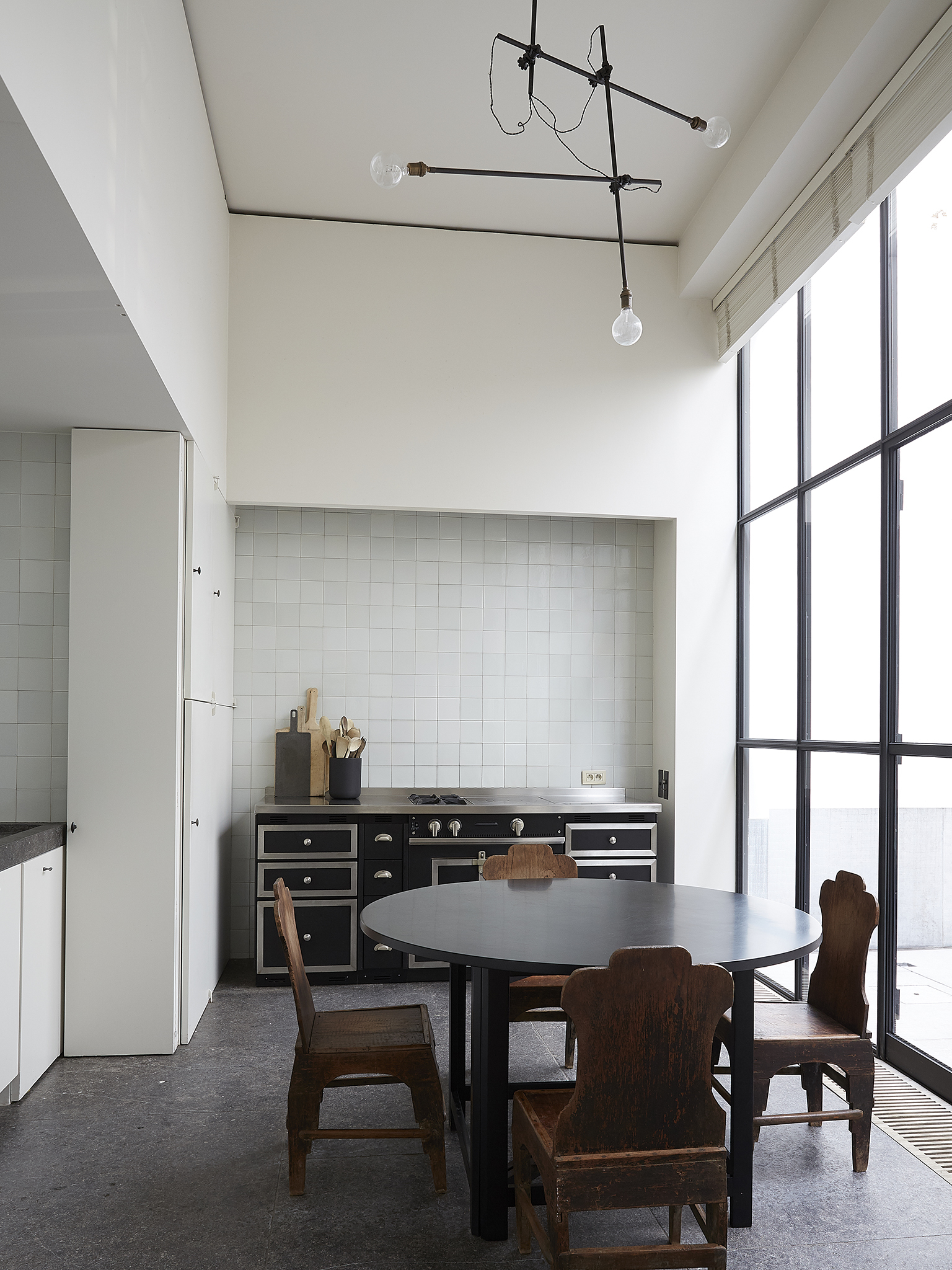 A round black dining table stands before a large steel-framed window, with carved wood chairs, a tiled alcove, and the black La Cornue range behind.