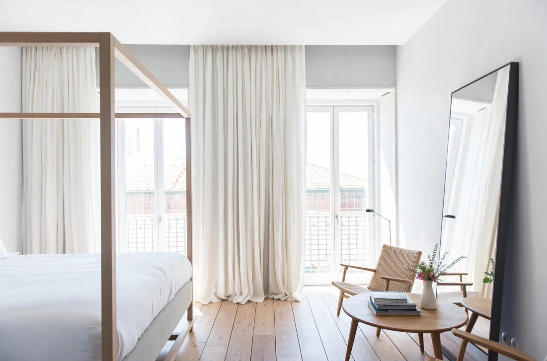 At the window wall, a pale timber canopy bed, floor-to-ceiling curtains, and a leaning black-framed mirror share the room with woven chairs and round wood tables.