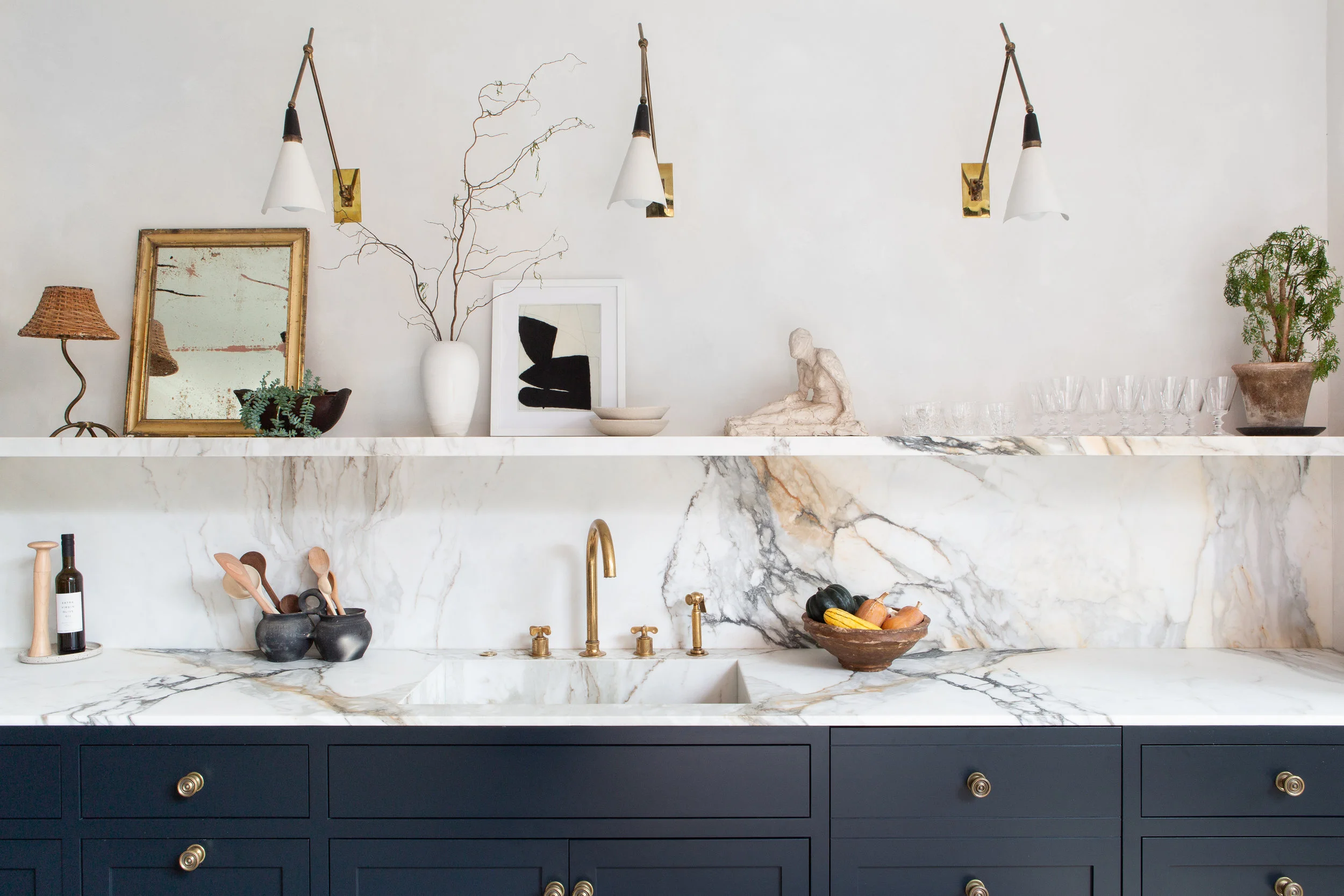 A tighter kitchen elevation highlights the veined marble sink run, brass tapware, blue painted drawer fronts, and a long marble shelf styled below brass-and-white articulated sconces.