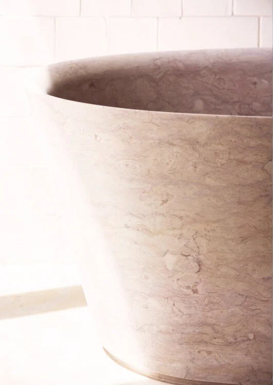 Sunlight rakes across an oval freestanding stone tub with soft brown veining, set against white rectangular wall tile.