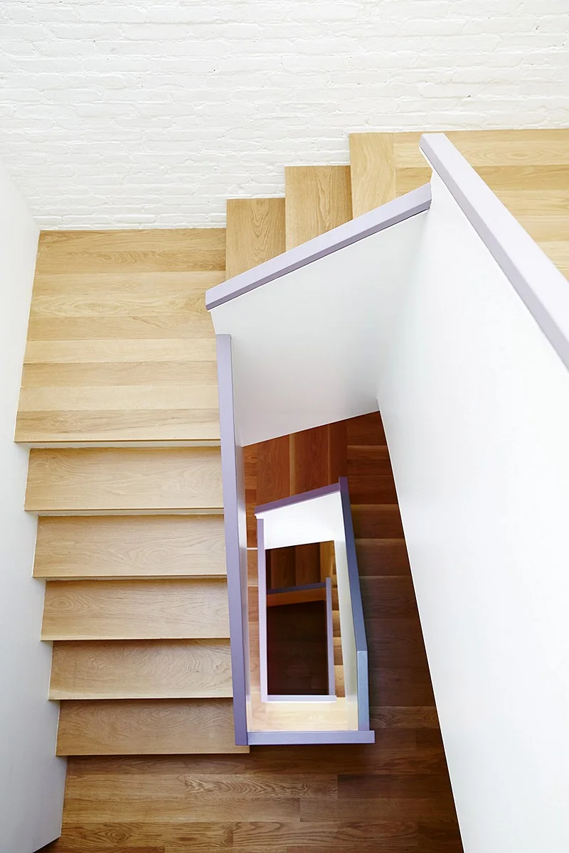 A switchback stair with pale oak treads and lavender-edged rails cuts through white brick walls.