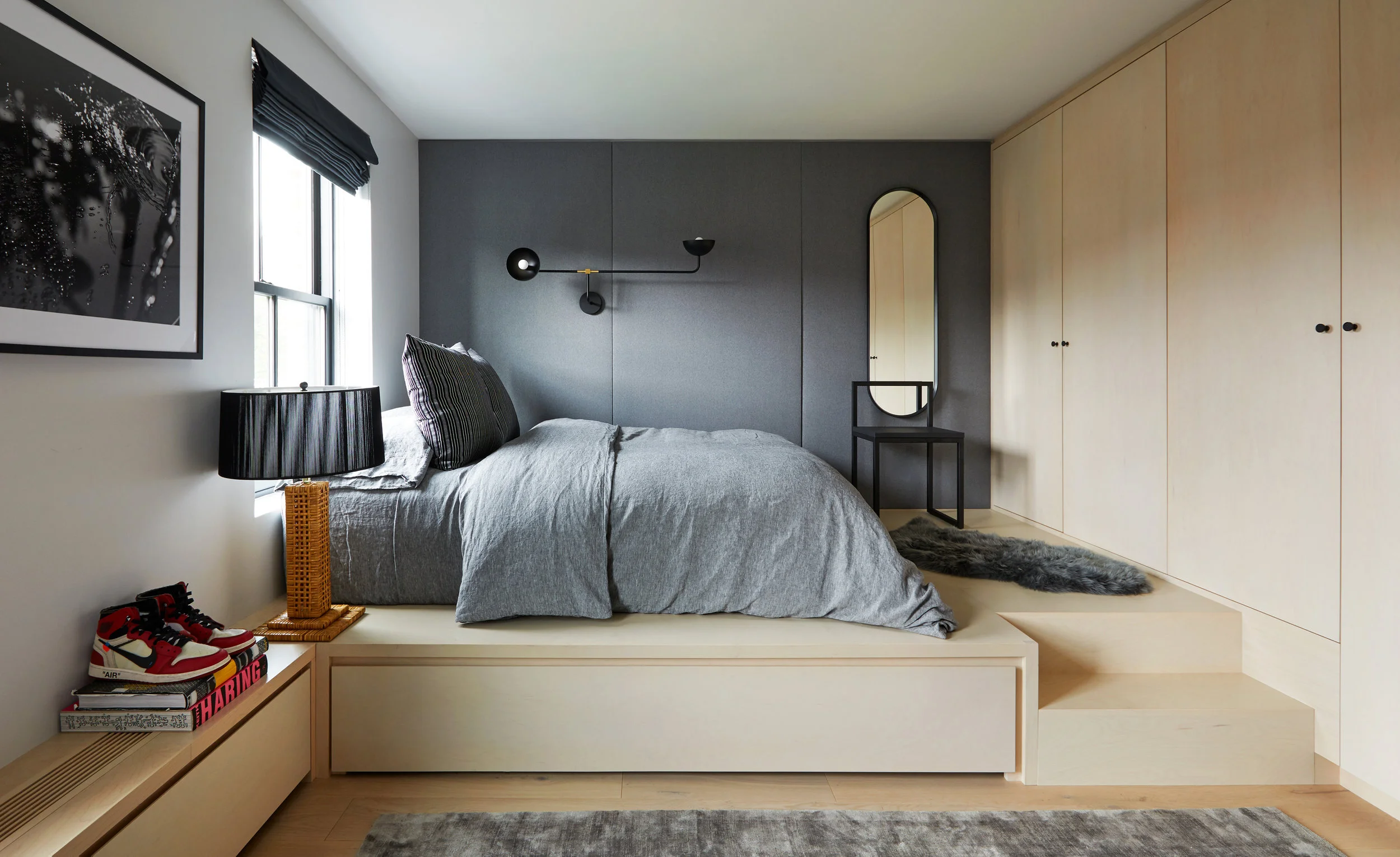 A raised platform bed, pale maple-toned built-ins, and a charcoal wall panel backdrop define this bedroom, with integrated steps and wardrobes turning the room into a continuous piece of millwork.