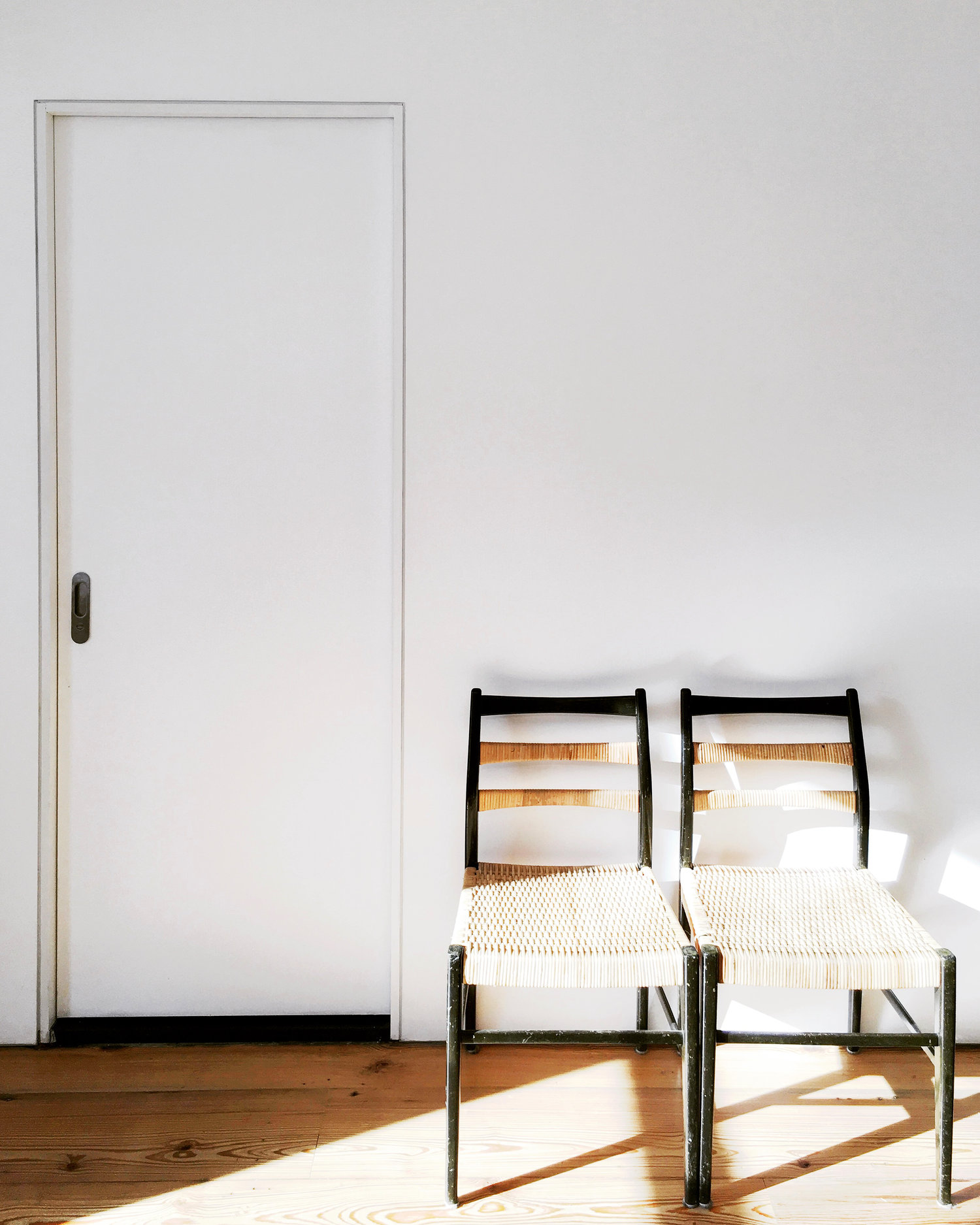 Two black-framed rush-seat chairs sit against a white wall and flush door, with sharp afternoon light falling across the wood floor.