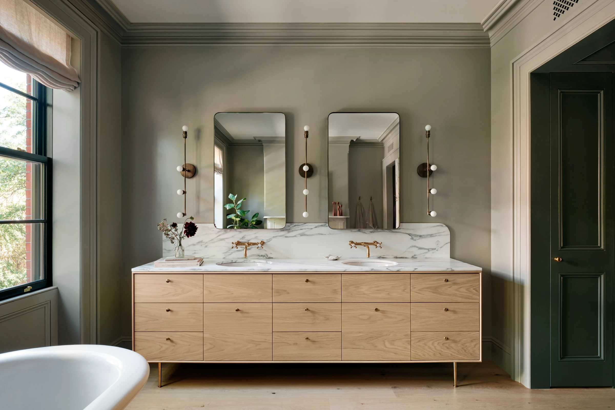 A curved oak vanity on brass legs sits against waxed plaster and white marble, showing the project's mid-century inflected detail control.