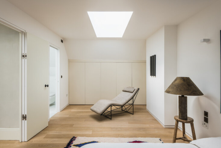 At the top-floor guest suite, a single rooflight brightens low built-in storage, wide-plank wood flooring, and a black-frame chaise lounge.