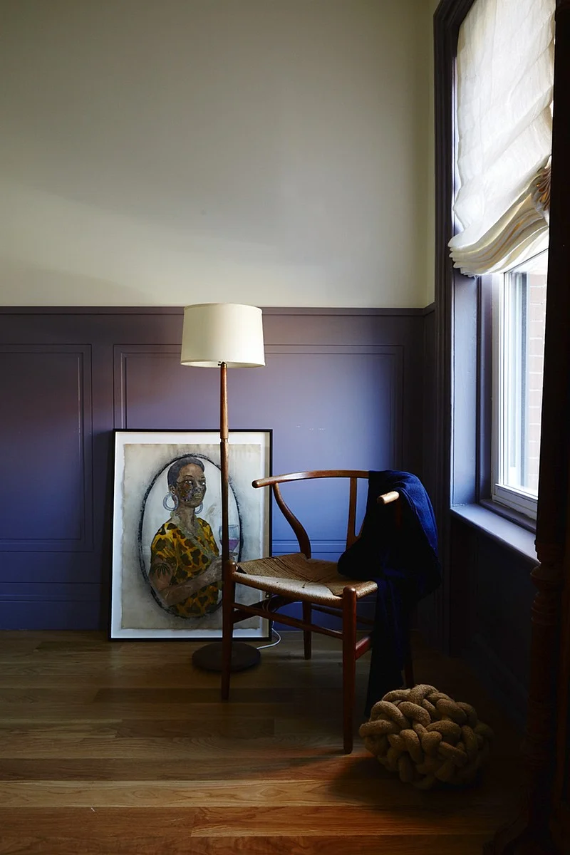 A corner vignette layers a cane chair, floor lamp, and portrait against deep lavender wall paneling.