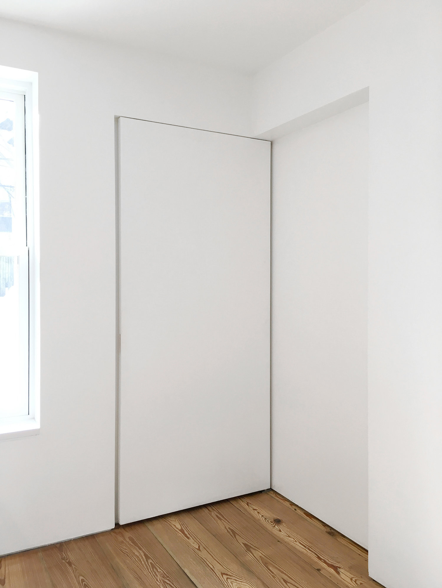 A flush white sliding door tucks into a wall pocket, meeting plain white walls and wide-plank wood flooring.