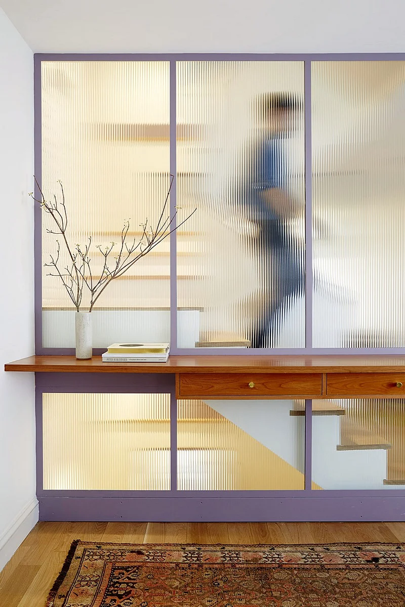 A ribbed-glass partition reveals a blurred figure moving on the stair beyond a slim console table.