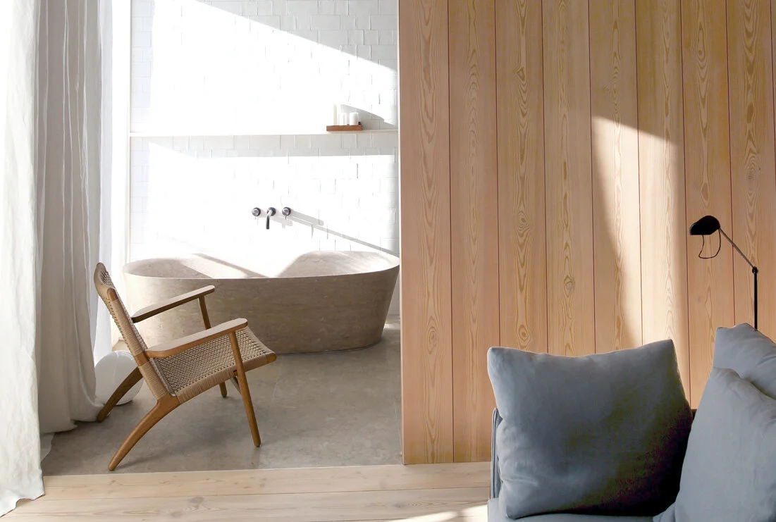 From sofa to bath, a cane lounge chair, wall-mounted taps, and a stone bathtub align across pale flooring beside vertical pine paneling.