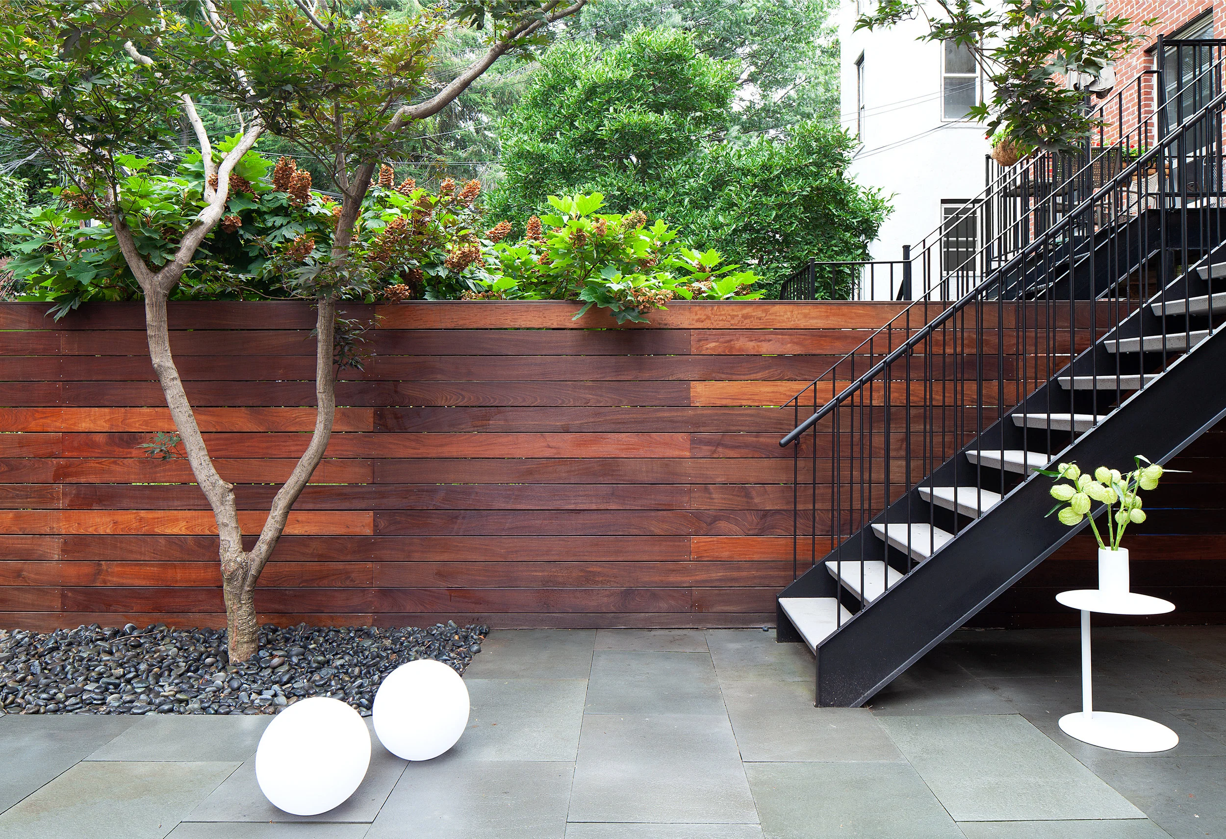 At garden level, black steel stairs meet dark-stained wood fencing above large-format stone pavers and white globe lights.