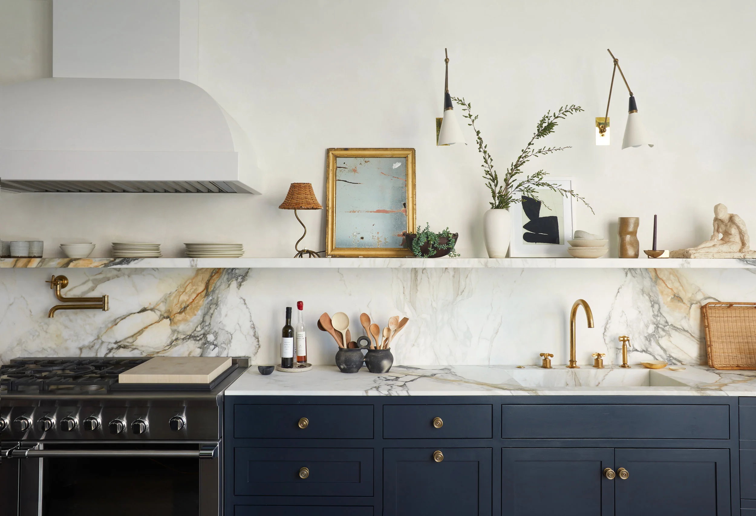 A white plaster hood and veined white marble shelf, splash, and countertop run across the kitchen wall above deep blue painted cabinetry, with brass pot filler and faucet fittings warming the palette.