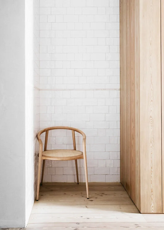 Set into a tiled recess, a woven-seat ash chair rests on light plank flooring beside white rectangular wall tile and a flush vertical timber panel.