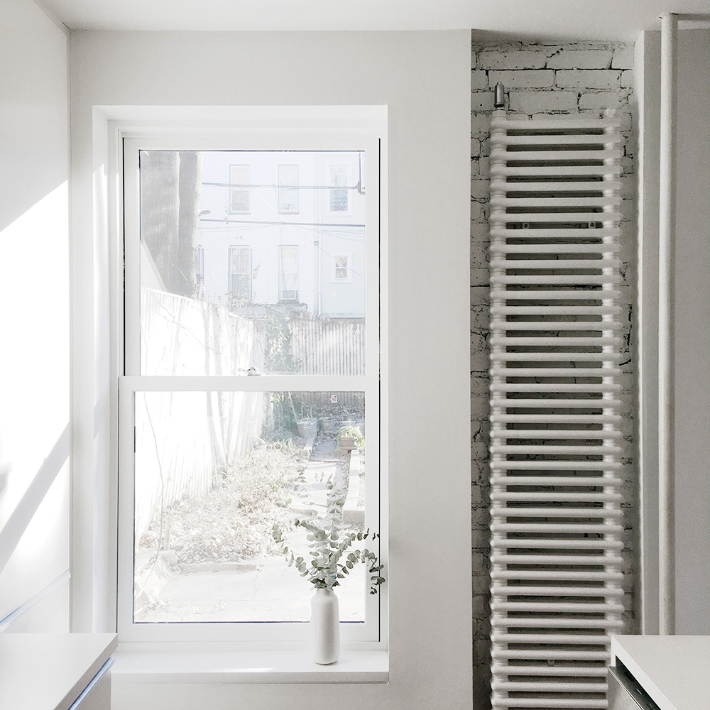 A tall rear window brings light into the white kitchen beside an exposed painted brick wall and vertical radiator.