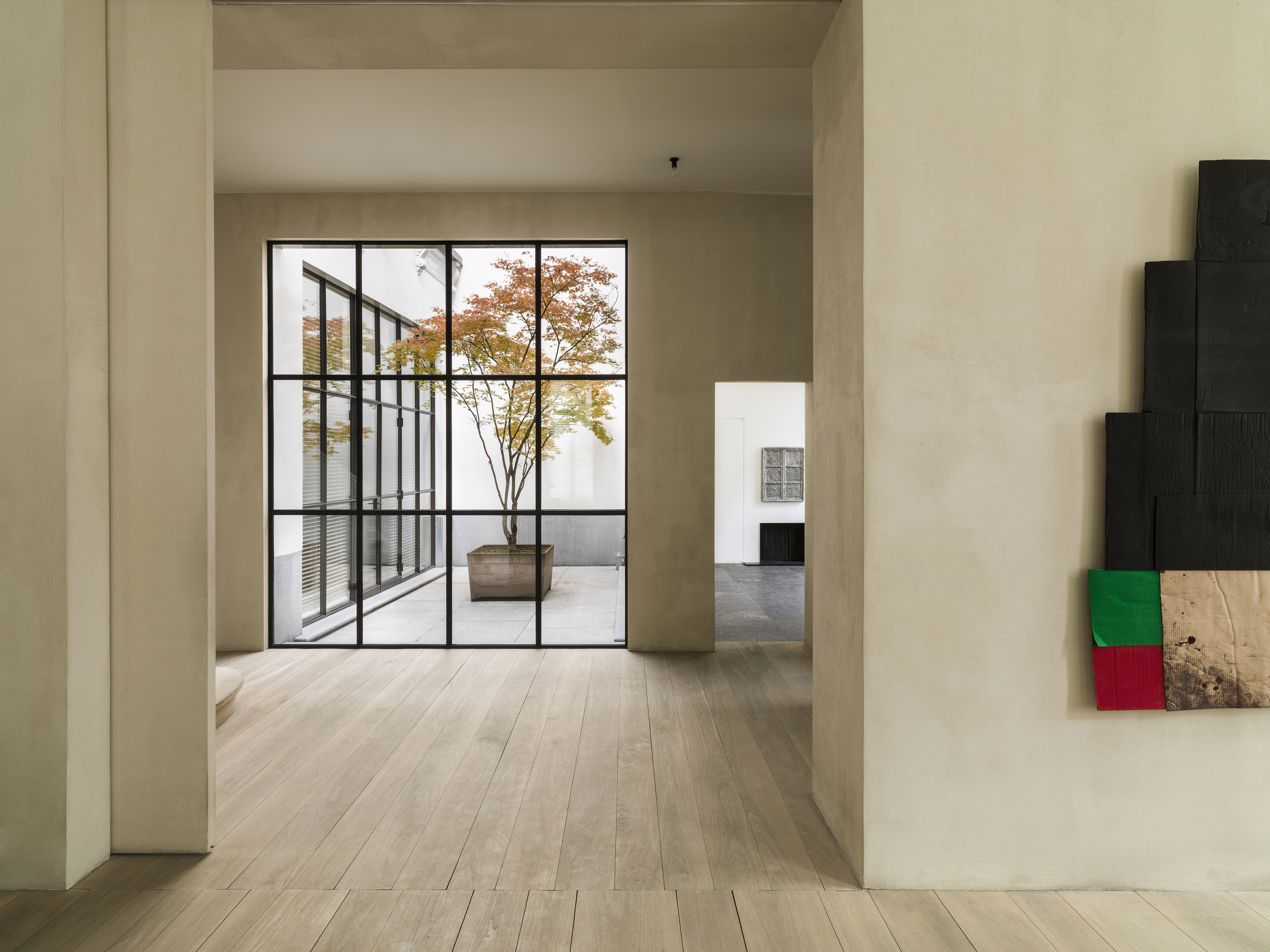 Black steel courtyard glazing frames a potted tree, while pale wide-board floors and thick plastered openings keep the adjoining rooms spare.