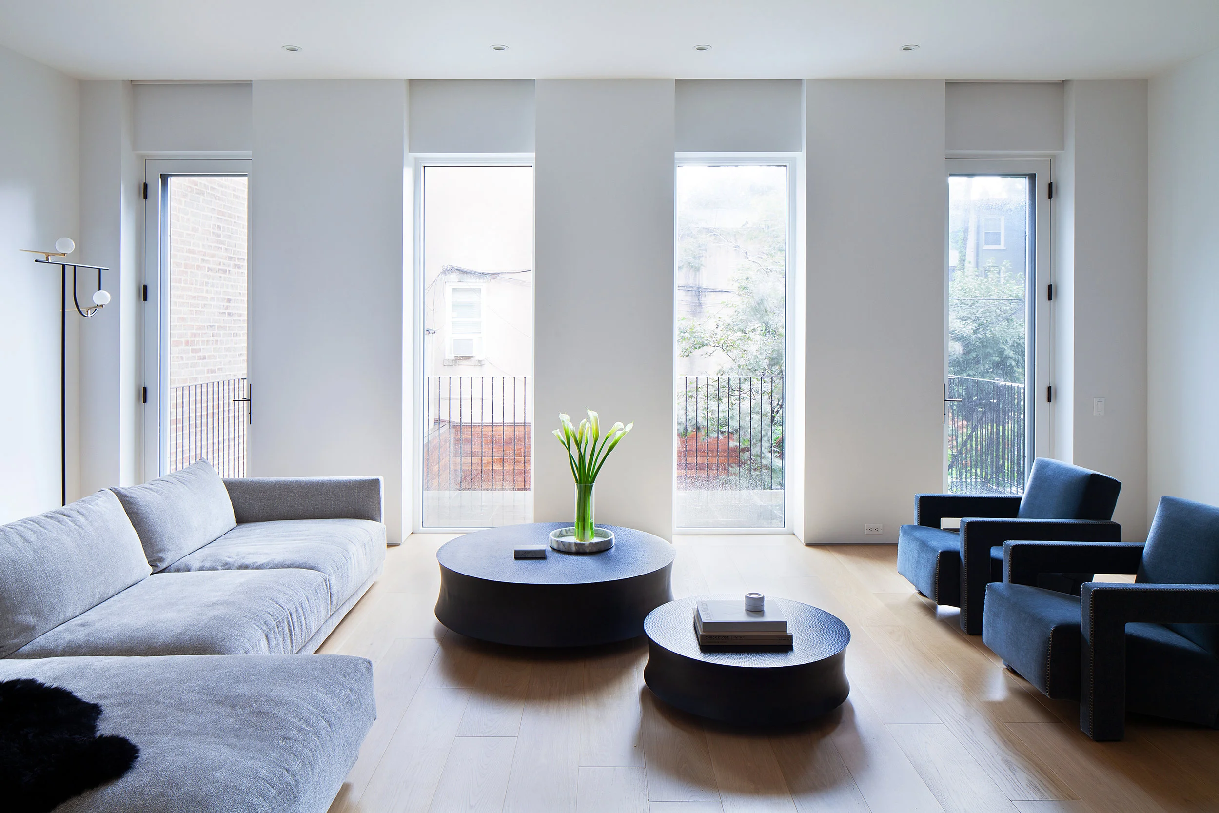 A long living room places a gray sectional and deep blue lounge chairs around two low black circular tables.