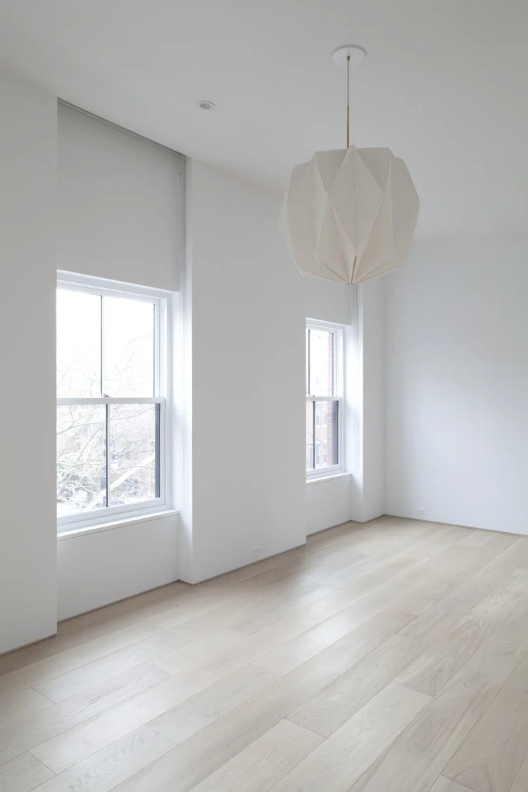 An upper room with two sash windows is finished with wide-plank white-oak flooring and a folded geometric pendant.