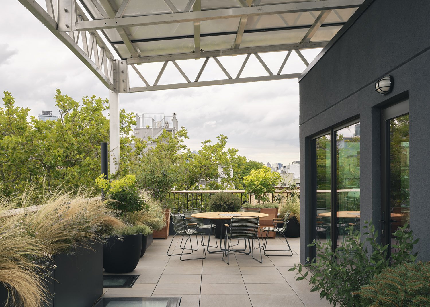 A steel canopy shades the roof terrace, where pale pavers, black wire chairs, and deep planters of grasses and shrubs frame the tree canopy beyond.