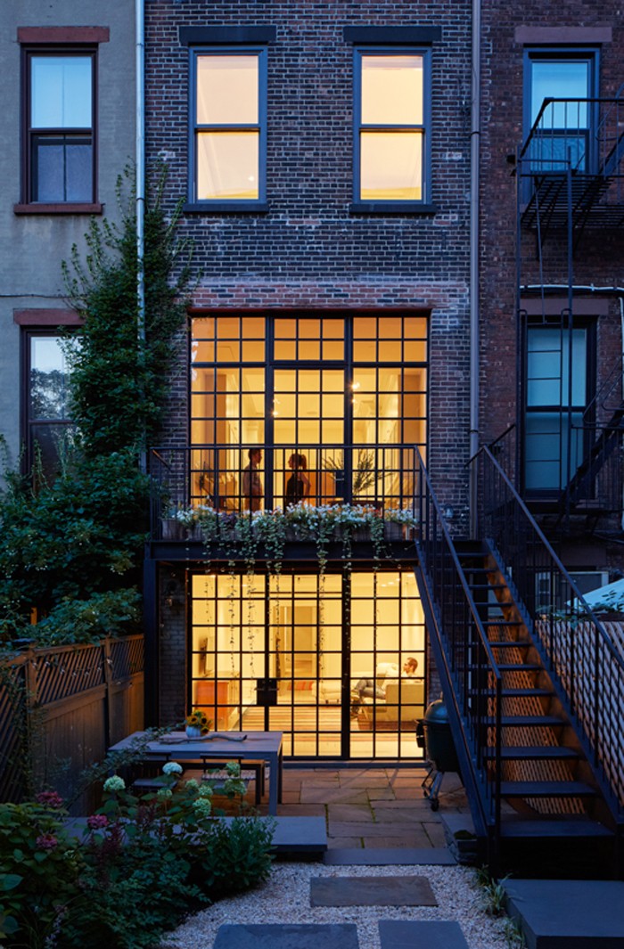At dusk, the rear facade glows behind black steel windows, showing stacked interior rooms and a planted garden path in front.