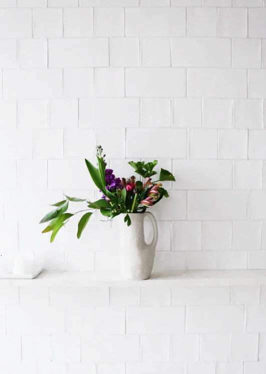 On a slim white ledge, a white ceramic jug with green and magenta stems sits against white stacked wall tiles.