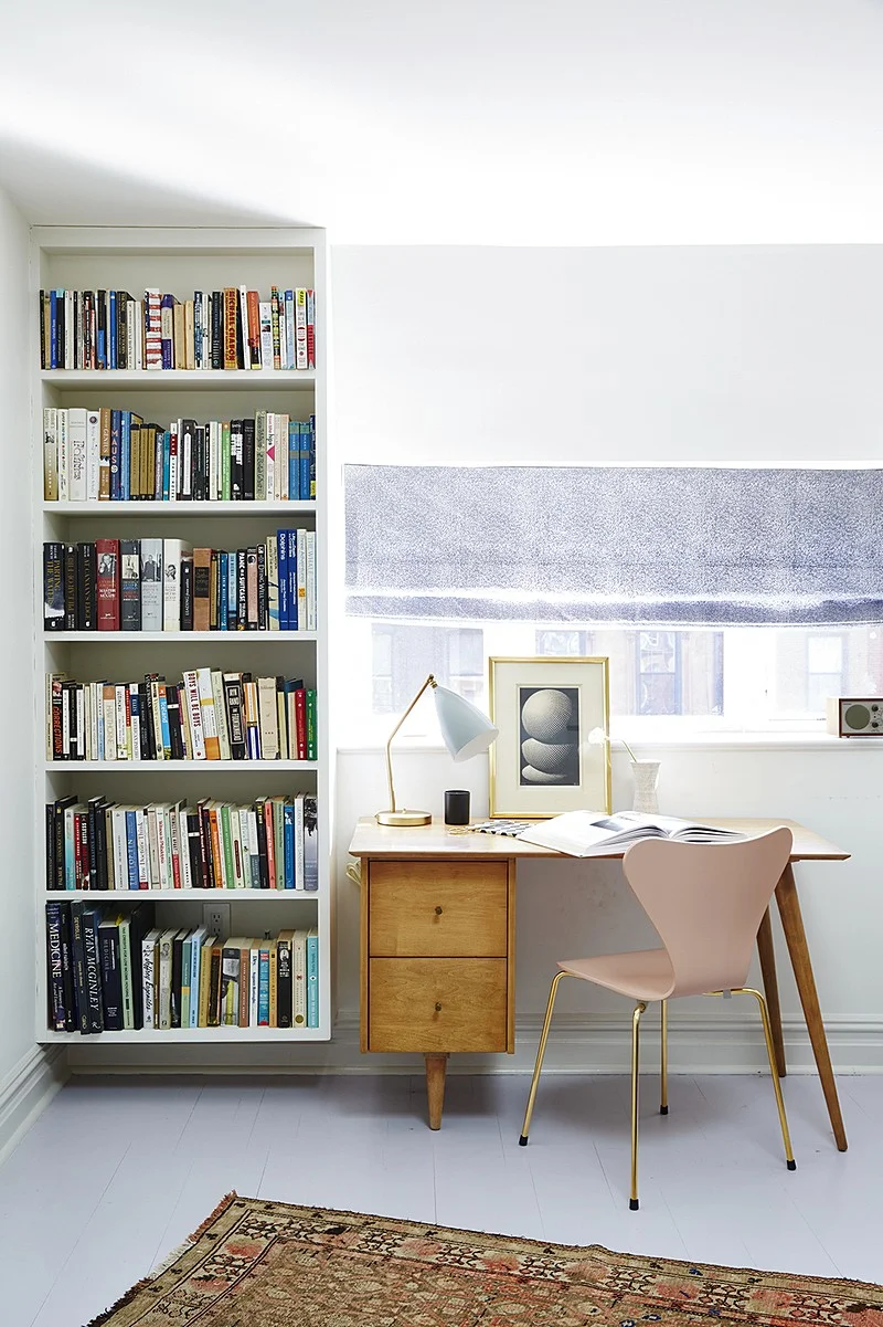 A compact study combines a tall bookcase, a timber desk, and a pale pink chair under a roman shade.