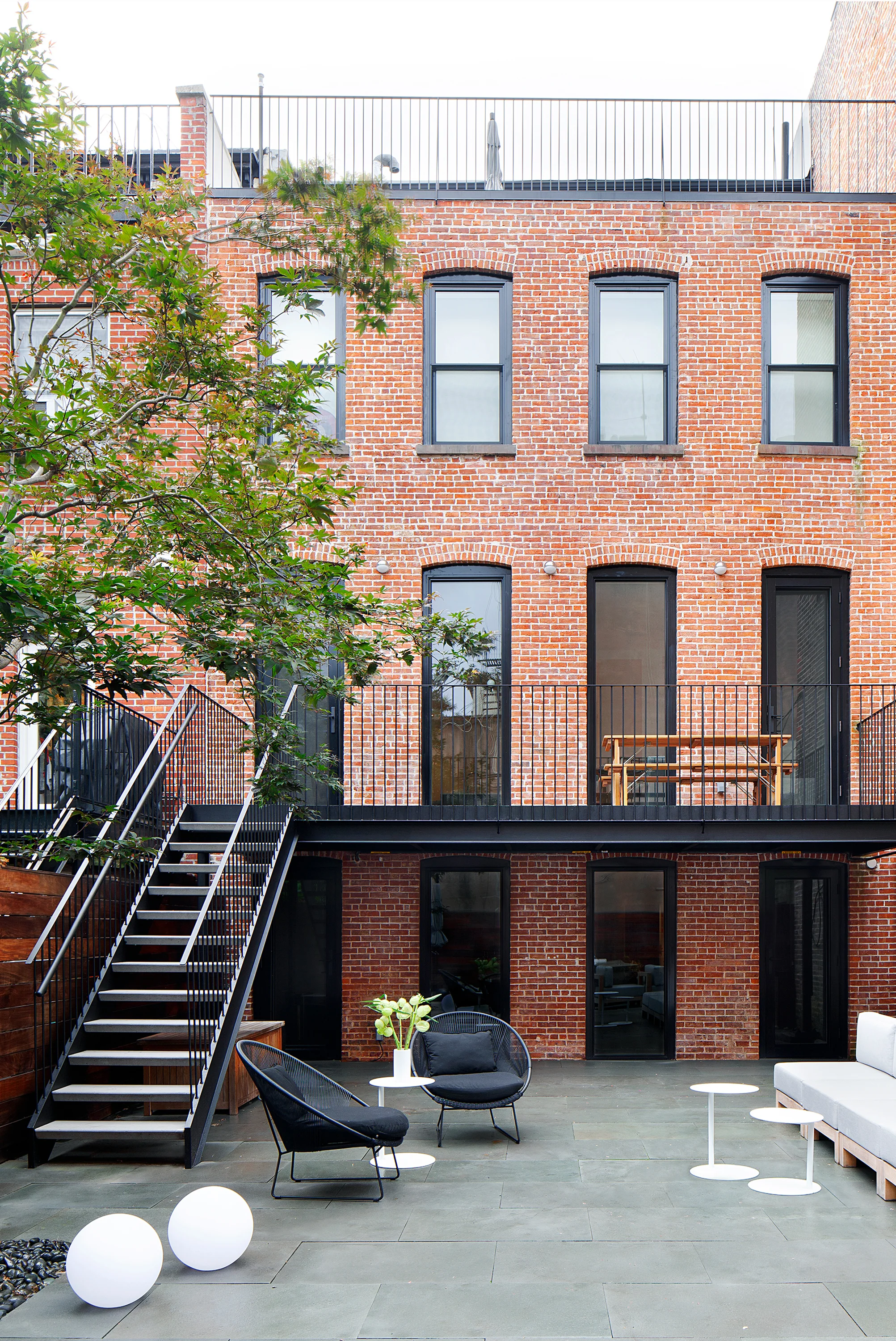 The rear elevation opens onto a paved terrace with black steel stairs, dark-stained wood fencing, and layered planting.