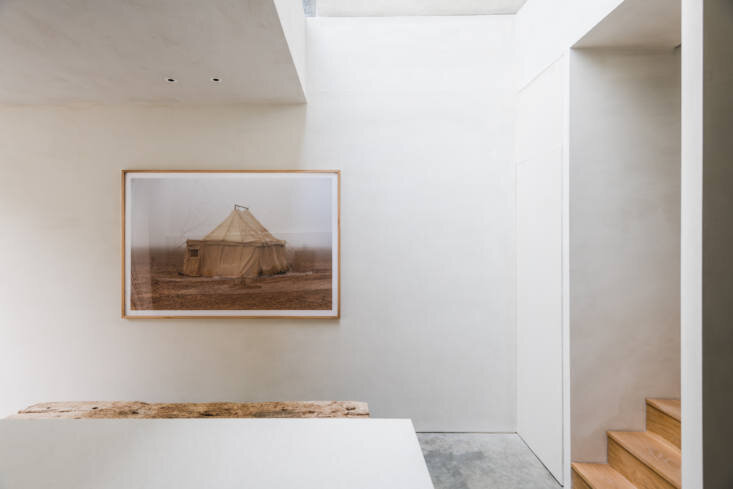 Light from the overhead well washes pale plaster walls, a framed artwork, and the tight stair turn beside the concrete floor.