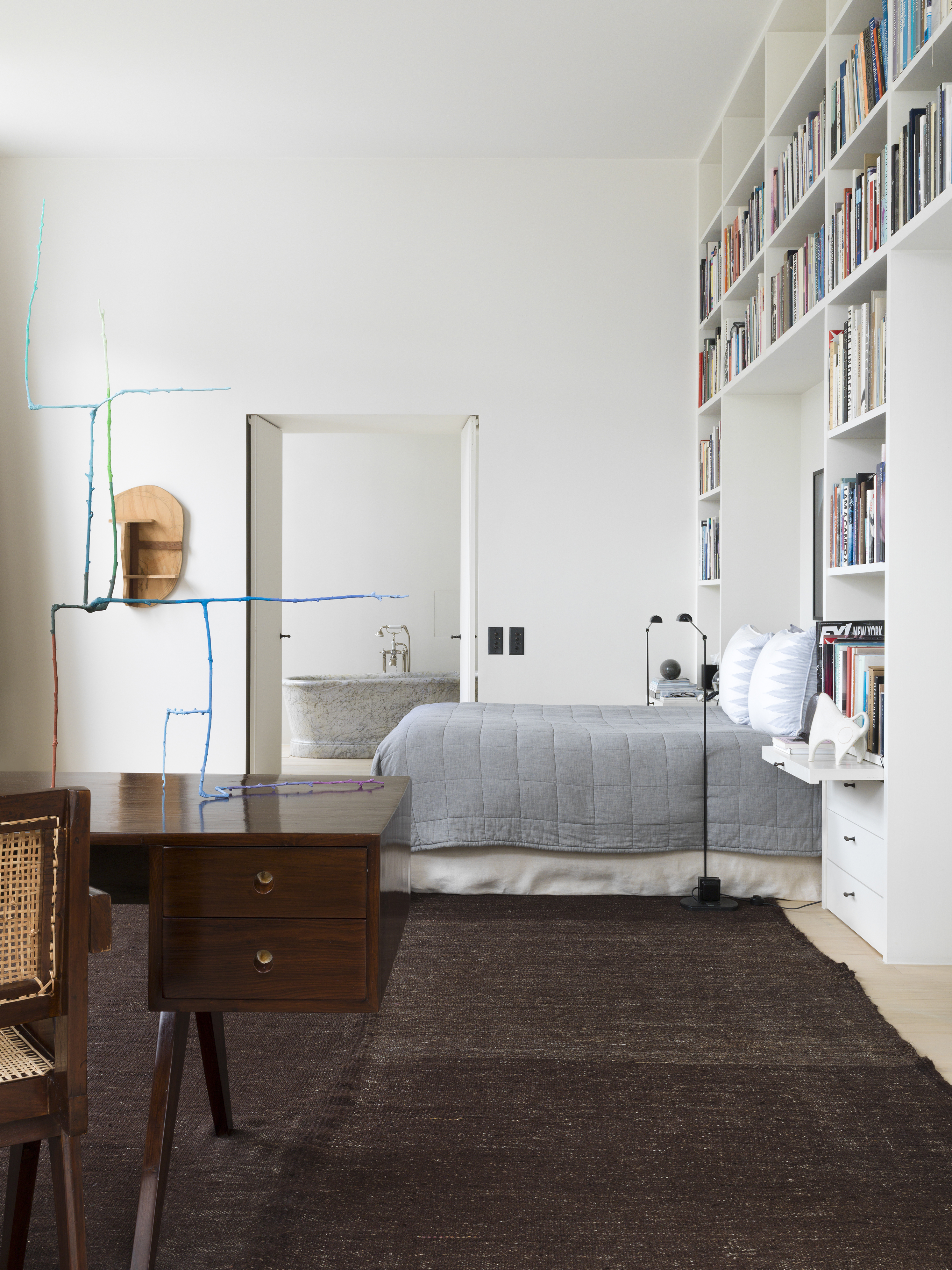 Built-in white bookshelves run above and beside a gray-covered bed, while a low doorway opens directly to the stone bath beyond.