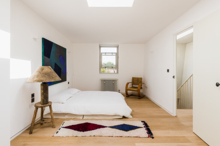 Guest room remains pared back with a floor-level bed, wide-plank wood flooring, a rooflight, and a woven lounge chair.
