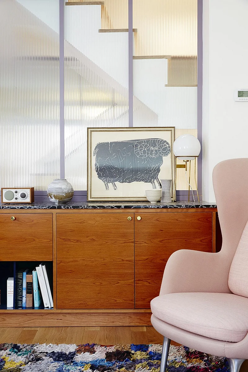 A walnut sideboard and pink lounge chair sit in front of a ribbed-glass partition with lavender framing.