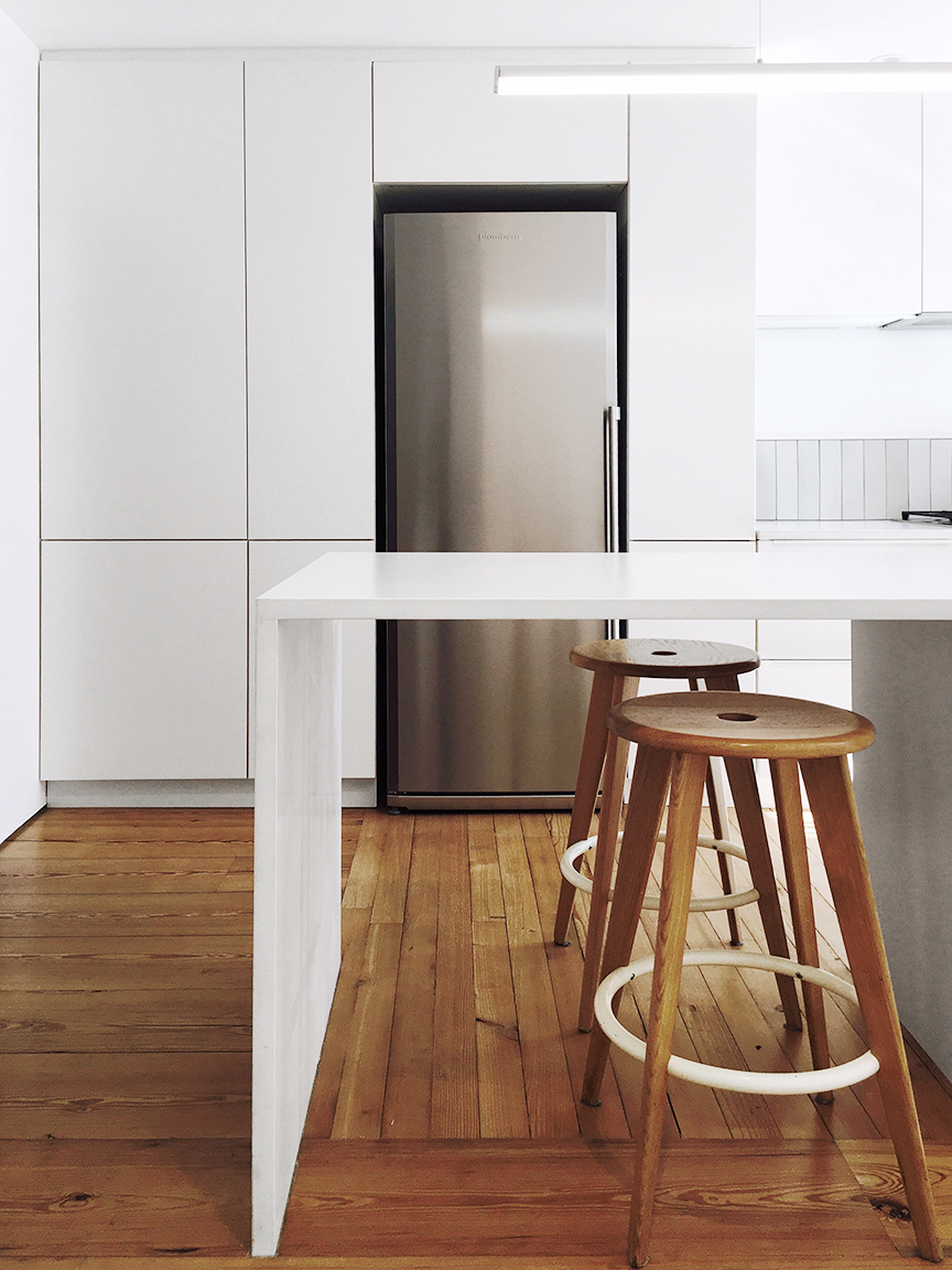 At the island edge, white cabinetry meets warm wood floors and two wooden stools beside the refrigerator wall.