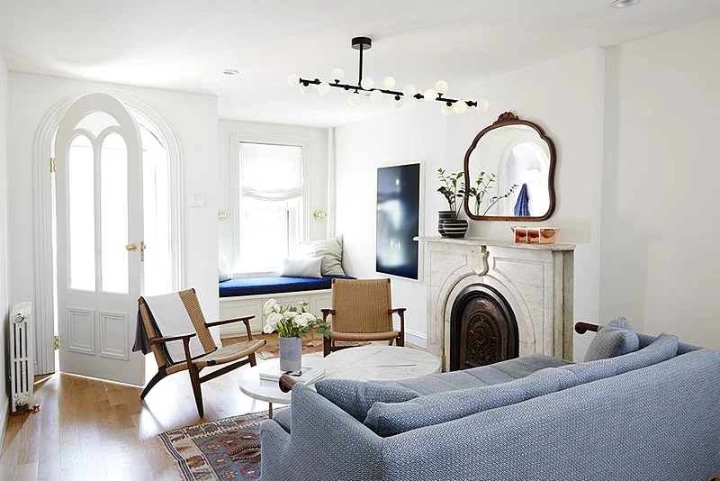 A bright sitting room pairs a marble fireplace with a blue sofa, woven chairs, and a window seat.