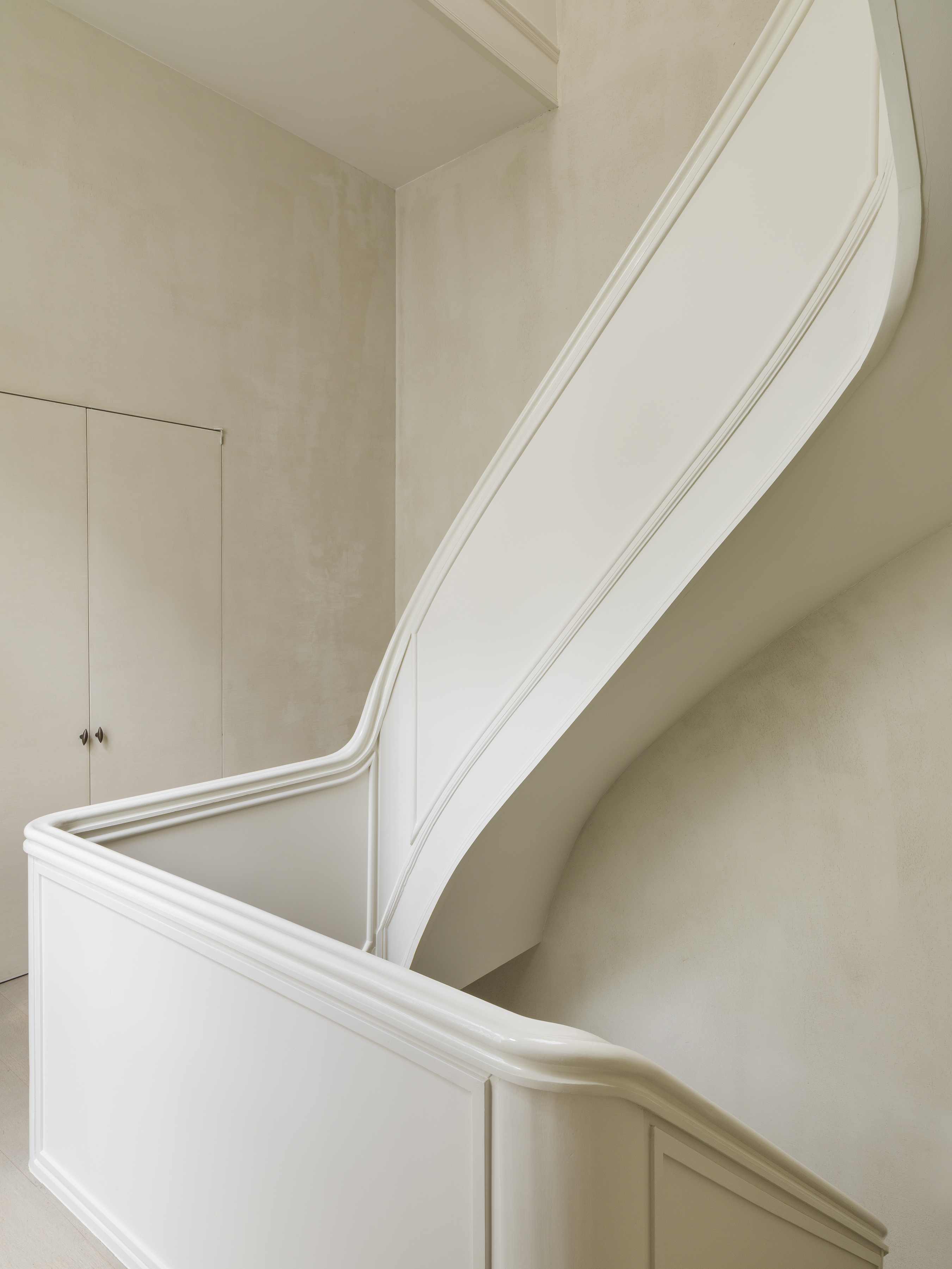 The stair balustrade is reduced to smooth white planes and rounded moldings, set against pale plaster walls and flush cupboard doors.