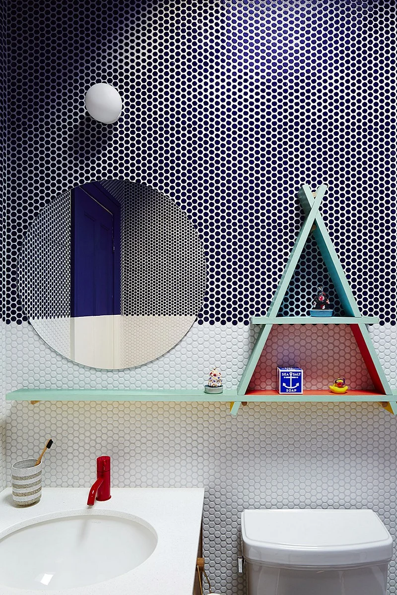 Navy-and-white penny tile wraps this bath wall with a round mirror, red faucet, and triangular shelf.