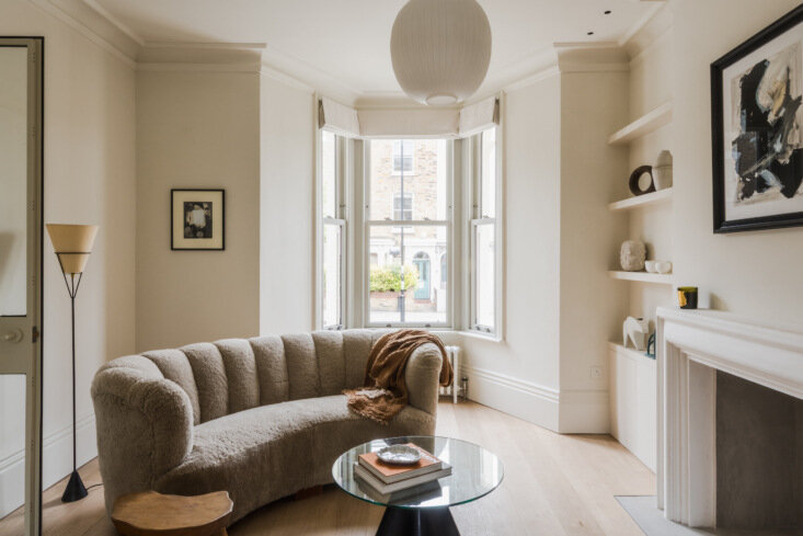 Curved 1940s sofa reupholstered in sheepskin wraps a round glass coffee table in the bright bay-window living room.