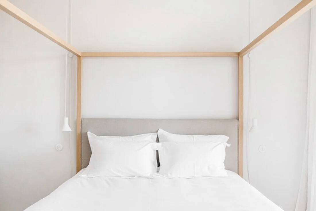 Within a pale alcove, crisp white bedding and a light upholstered headboard sit under a simple blond-wood canopy with white pendant reading lights.