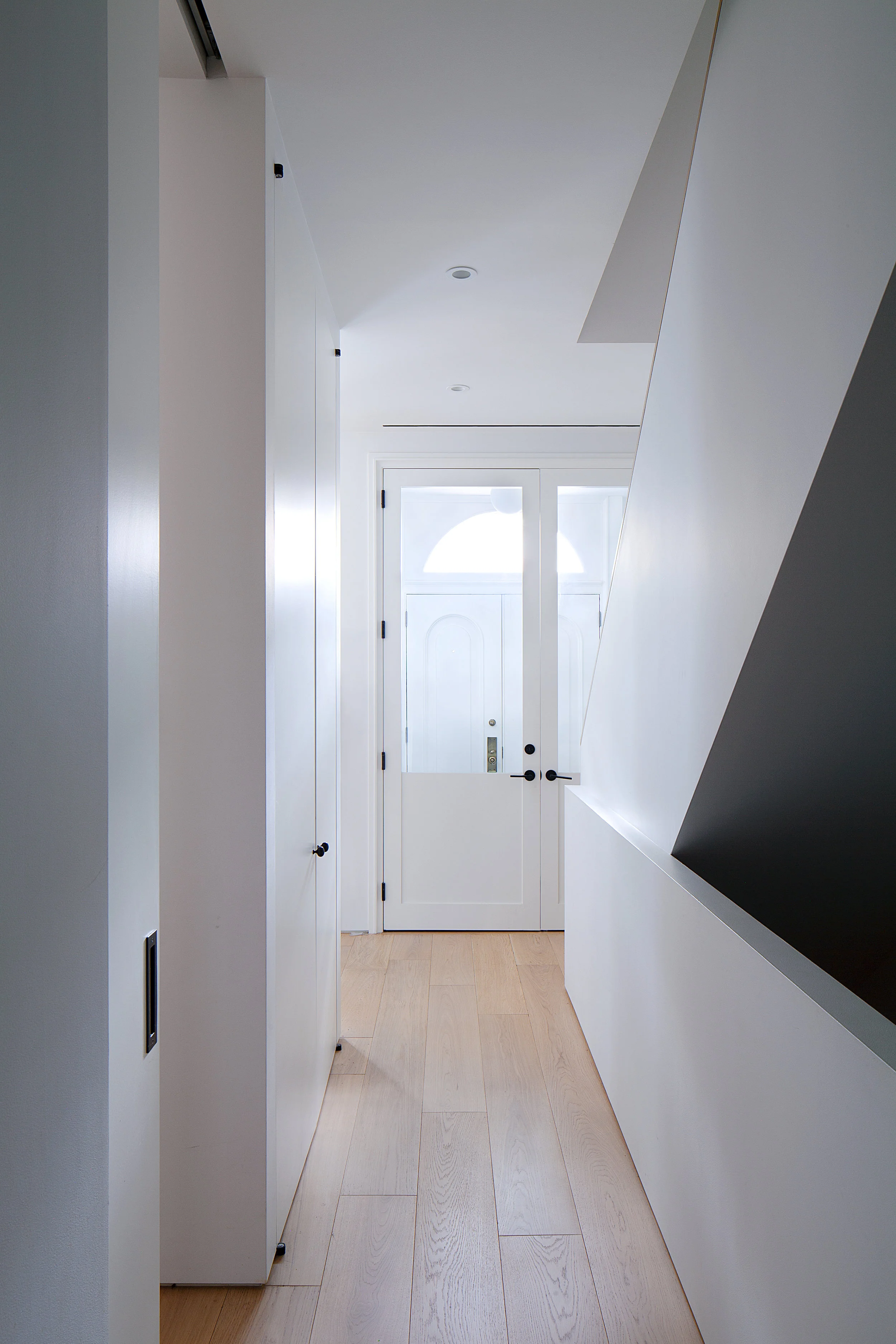 A narrow hall uses wide-plank white-oak flooring, flush white walls, and a sharply angled black stair enclosure.