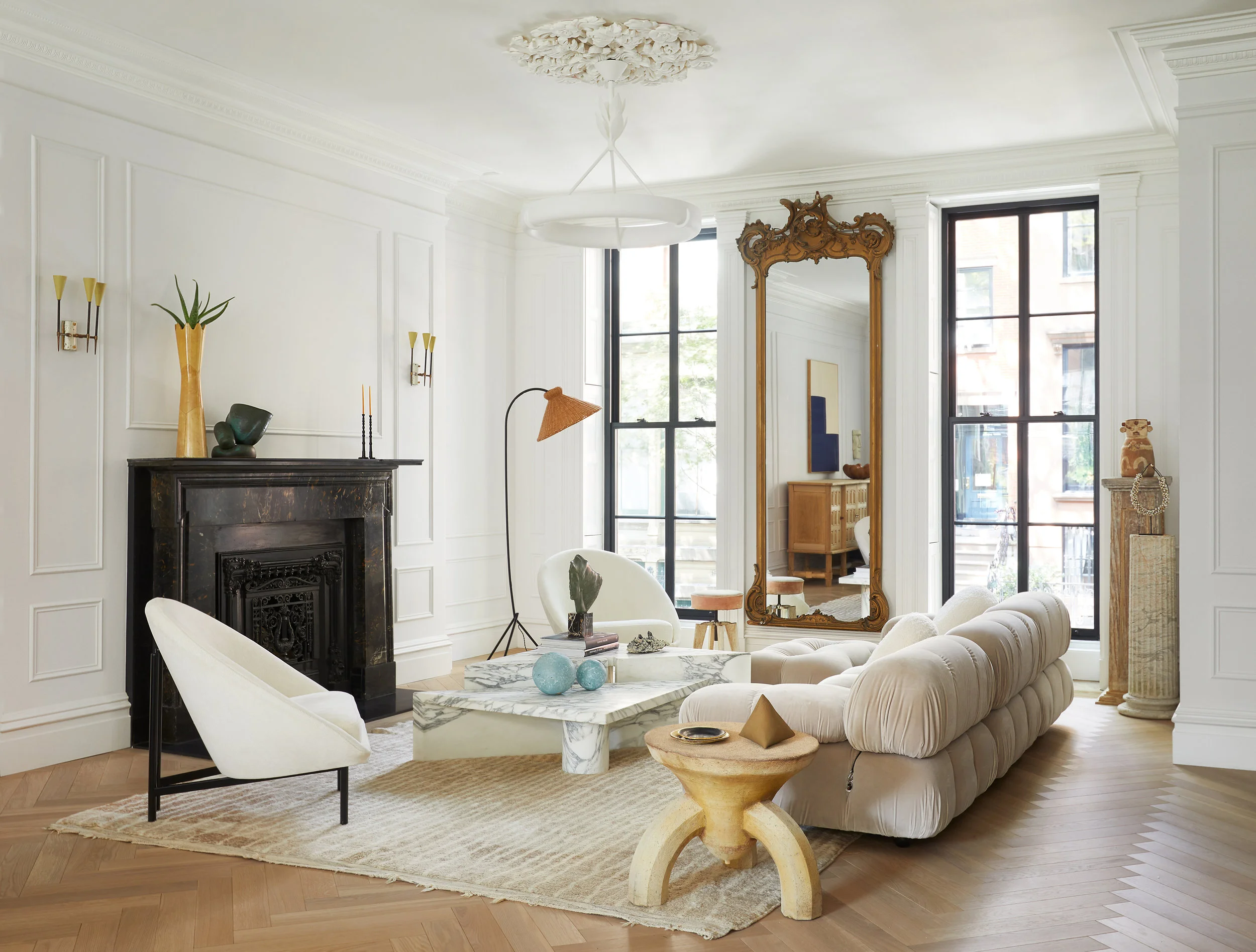 In the formal living room, a black stone fireplace anchors white paneled walls, while a channel-tufted cream sofa, white lounge chairs, a veined marble coffee table, and a tall gilt mirror layer old and new elements.