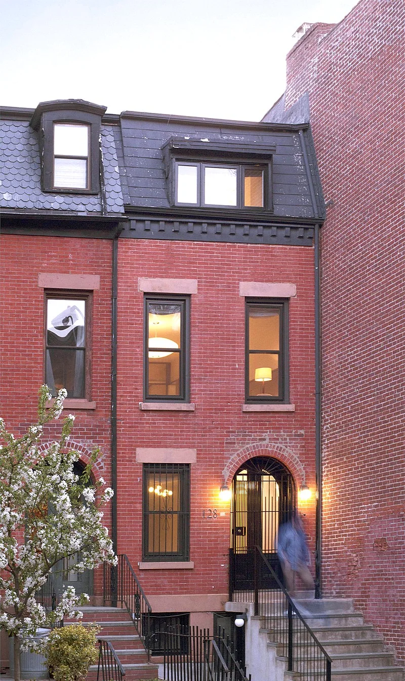 At dusk, the red-brick townhouse facade shows lit windows, a black mansard roof, and a glowing entry.