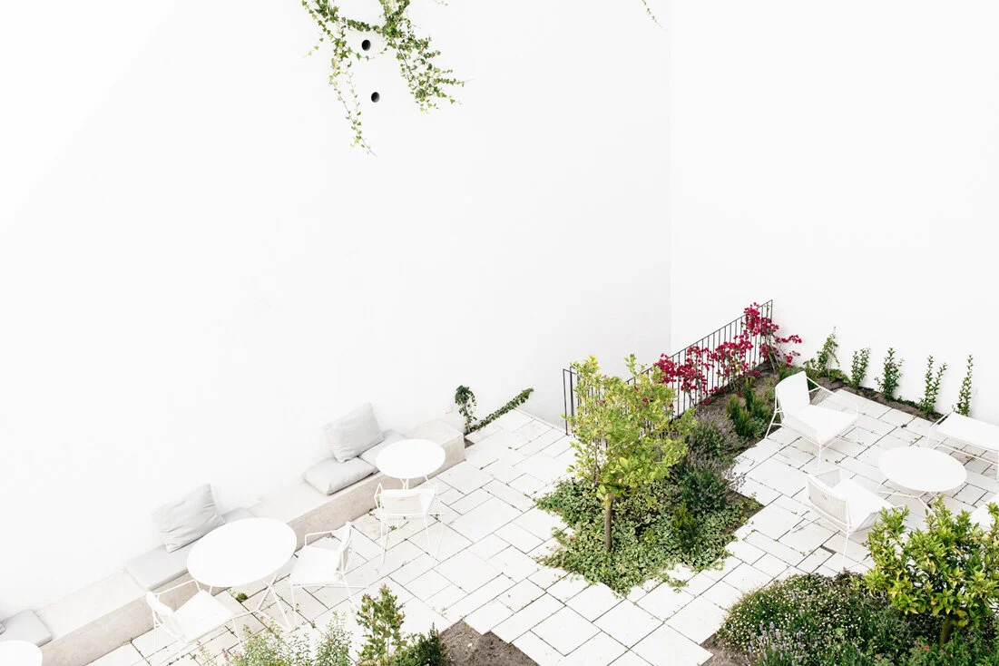 Viewed from above, white paving slabs, round white tables, and wire chairs wrap planted beds while pink vines climb a black stair rail.