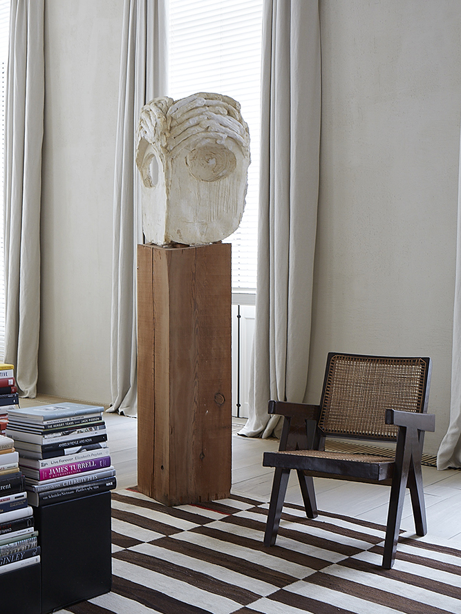 A striped flatweave rug, a dark wood chair with a cane seat, and a plaster head on a timber pedestal sit between tall drapes and pale walls.