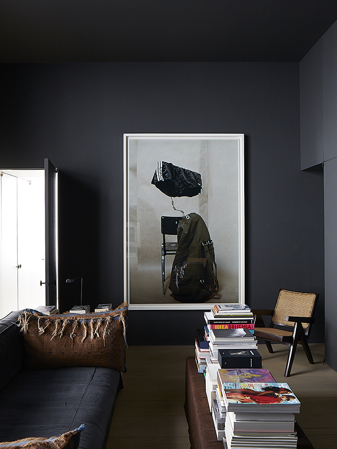 Dark charcoal walls set off a large framed artwork, a black sofa, a cane chair, and stacks of books on a long brown bench.