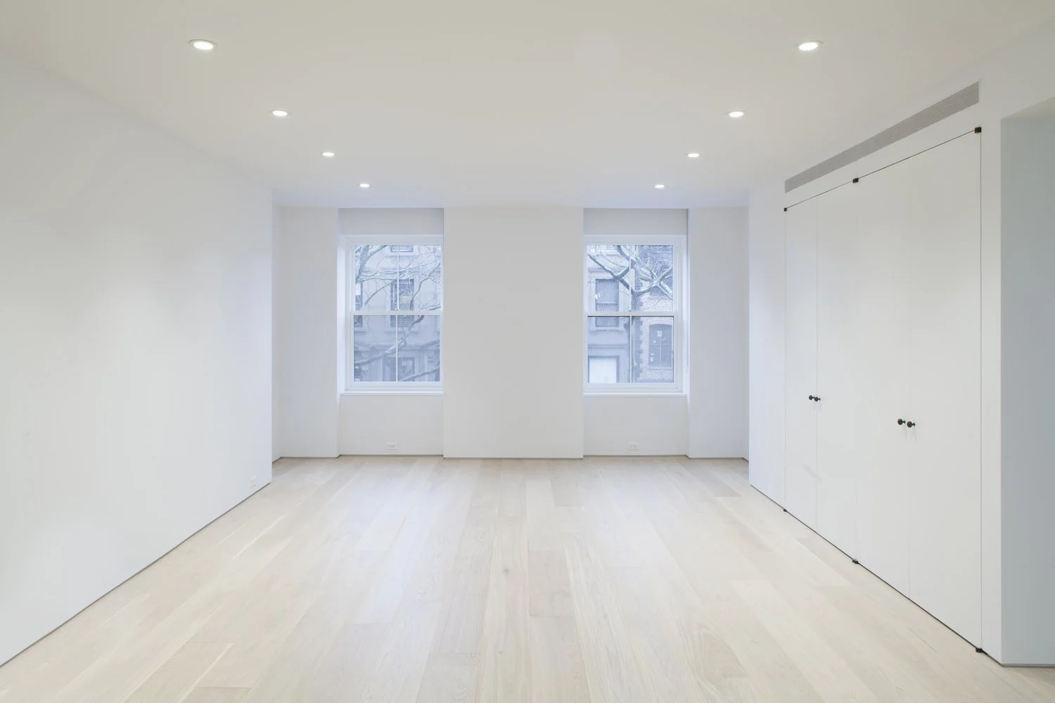 A large white room with two front-facing windows is kept spare by flush full-height storage and recessed ceiling lights.