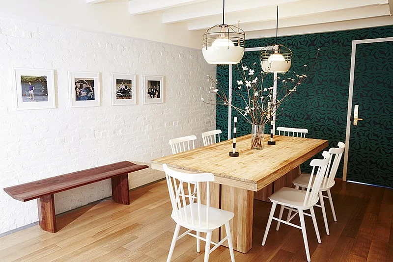A long timber dining table and bench are set against white-painted brick and dark botanical wallcovering.