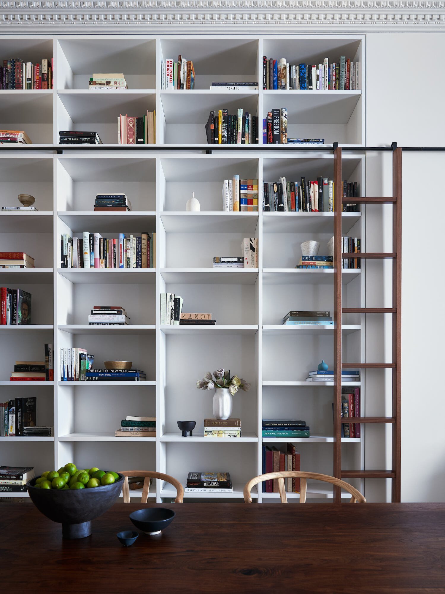 A full-height white bookcase with a sliding wood ladder forms the dining backdrop, with a dark wood tabletop and curved dining chairs in the foreground.